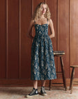 A woman with long blonde hair stands indoors against wood paneling, wearing The Verse Dress by The Great Inc.—a blue cotton dress with a floral pattern and sculpted silhouette—paired with black high-top sneakers. She rests a hand on books atop a wooden stool.
