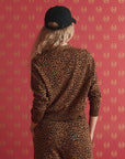 A person with long, wavy blonde hair, wearing a black cap and The Great Inc. Shrunken Vneck Cardigan, stands facing a red patterned wall, shown from behind.
