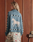 A person with wavy blonde hair, seen from behind, wears The Stable Lodge Cardigan by The Great Inc., featuring a bold western horse motif and patterned sleeves, paired with a floral skirt in front of wooden paneling.