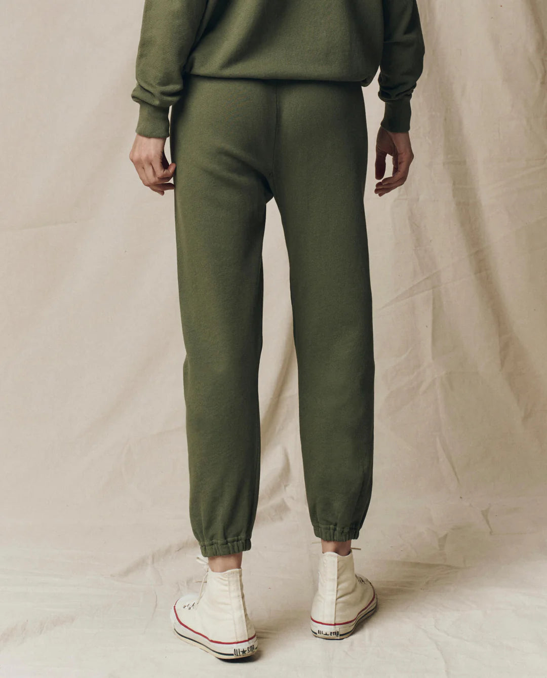 Wearing The Great Inc.'s handcrafted, locally made olive green Stadium Sweatpant and matching sweatshirt with white high-top sneakers, a person stands on a beige fabric backdrop facing away from the camera.