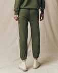 Wearing The Great Inc.'s handcrafted, locally made olive green Stadium Sweatpant and matching sweatshirt with white high-top sneakers, a person stands on a beige fabric backdrop facing away from the camera.