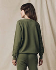 A woman with long brown hair wears The Great Inc.'s oversized College Sweatshirt S25 and matching pants, standing with her back to the camera against a beige fabric backdrop.