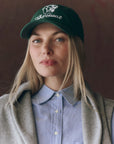 A woman with long blonde hair wears The Baseball Hat by The Great Inc., featuring vintage chainstitch embroidery of "The Great" and a tiger, along with a light blue button-up shirt and gray coat, against a dark background.