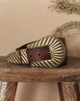 The Big Buckle Belt by The Great Inc., made from top-grain leather with a large fluted buckle featuring textured leaf-like patterns, is coiled on a rustic wooden stool against a beige background with dried grasses.