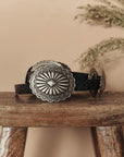 The Great Inc. Leather Concho Belt, crafted from top-grain leather with large, ornate silver conchos, sits on a rustic wooden stool with dried greenery and a beige wall in the background—showcasing true Western craftsmanship.