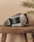 The Big Buckle Belt by The Great Inc.—a black top-grain leather belt with a large, decorative silver buckle—rests coiled on a rustic wooden stool. Dried foliage appears blurred in the background.