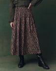 Wearing The Great Inc.’s The Canary Skirt—an airy pull-on style with red and yellow floral pleats—paired with a dark green jacket and black lace-up boots, person stands against a matching background, lower half only visible.