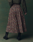 Someone wears The Canary Skirt by The Great Inc., an ankle-length cotton seersucker skirt with red and beige floral designs, paired with black boots and a dark green jacket, standing against a matching dark green background.