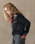 A woman with wavy blonde hair wears The Heirloom Rose Western Shirt by The Great Inc.—a black top with rose embroidery and pearl snaps—paired with a red bandana and gray jeans, standing in profile against a beige background, hands in her pockets.