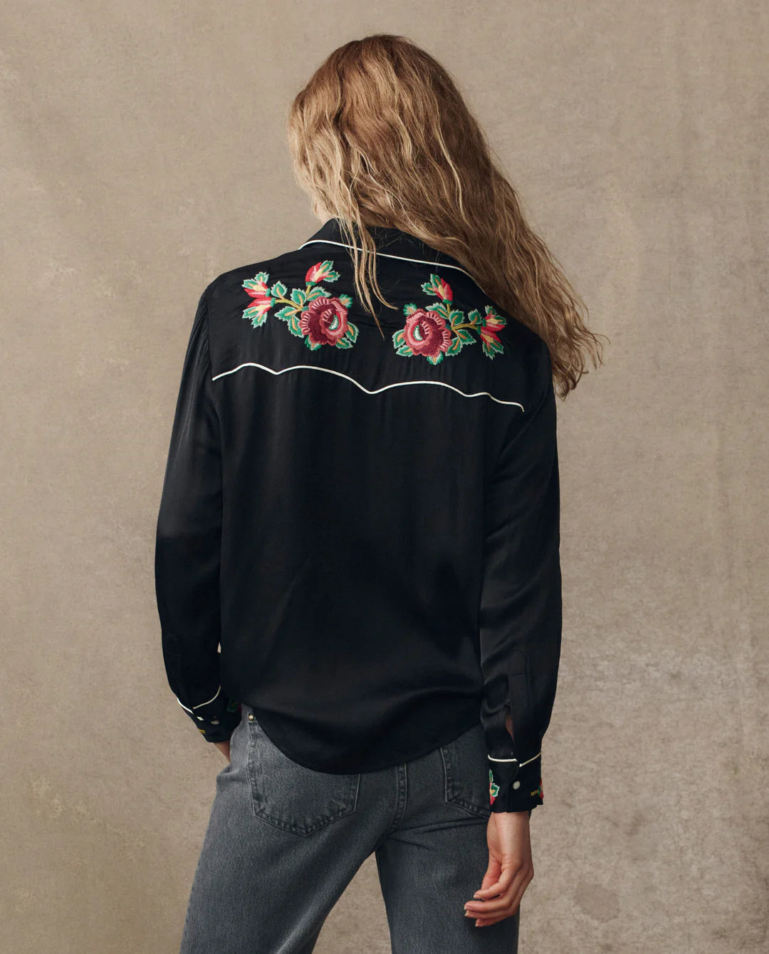 Seen from behind, a person with long wavy hair wears The Heirloom Rose Western Shirt by The Great Inc., featuring pearl snaps and red and pink rose embroidery on the upper back, paired with gray jeans against a plain background.