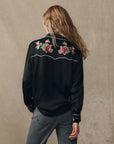 Seen from behind, a person with long wavy hair wears The Heirloom Rose Western Shirt by The Great Inc., featuring pearl snaps and red and pink rose embroidery on the upper back, paired with gray jeans against a plain background.