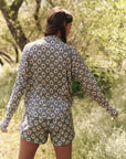 A person with long hair stands in a grassy area, facing away, wearing a long-sleeve shirt and The Rover Short by The Great Inc., featuring a blue and white floral print. Trees and greenery provide an ideal backdrop for showcasing practical outdoor apparel.