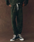 Wearing The Great Inc.'s The Stadium Sweatpant in dark green camouflage with a white drawstring and black high-top Converse sneakers, a person stands against a brown textured background; upper body not visible, evoking vintage athletic style.