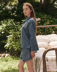 A woman with long hair stands outdoors by a vintage brass bed with white bedding, wearing The Pajama Shirt from The Great Inc. Lush green plants and flowers in the background create a dreamy atmosphere.