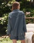 A person with long, light brown hair stands outdoors, back to camera, wearing The Pajama Shirt by The Great Inc. in blue with matching shorts. Greenery and a bed with pale bedding add vintage charm to the scene.
