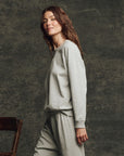 A woman with long brown hair wears The Fleece Rookie Sweatshirt by The Great Inc. in light gray, paired with matching sweatpants, standing sideways against a dark textured background. A wooden chair is partially visible on the left.