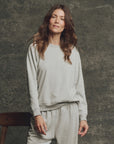A woman with long brown hair wears The Fleece Rookie Sweatshirt by The Great Inc., paired with matching sweatpants, as she stands smiling slightly before a textured gray backdrop.