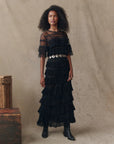 A woman poses before a beige backdrop wearing The Winter Mesh Flounce Skirt by The Great Inc., styled with a black dress featuring tiered ruffles, sheer sleeves, a wide silver belt, black boots, and stacked wooden crates beside her.
