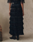 Only the lower body is visible as someone stands against a neutral, textured backdrop, wearing The Great Inc.'s The Winter Mesh Flounce Skirt— a long, black, mesh number with tiered ruffles—paired with black boots.