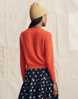 A person wearing a tan beanie, The Cashmere Tiny V-Neck Cardigan by The Great Inc. in orange, and a black skirt with white polka dots stands facing away from the camera against a neutral fabric background.