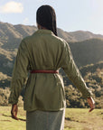Seen from behind, a person with long braids stands outdoors wearing The Vintage Army Jacket by The Great Inc. and a denim skirt, with green hills and a partly cloudy sky in the background.
