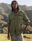 A woman with long braided hair stands outdoors before mountains, confidently wearing The Vintage Army Jacket by The Great Inc. in cotton slub twill and a light denim skirt, one hand in her pocket as she looks at the camera.