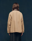 A person with long brown hair, wearing The Great Inc.'s The Barn Jacket with utility pockets and a dark skirt, stands facing away from the camera against a dark blue background.