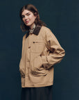 A person with long brown hair wears The Barn Jacket by The Great Inc., a tan workwear style with utility pockets and a dark brown collar, against a dark blue background.