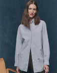 A woman with long brown hair models The Great Inc.'s Menswear Button Up, featuring subtle pinstripes, as she stands before a dark blue background near a wooden chair, smiling gently at the camera.