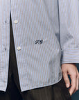 A close-up showcases The Menswear Button Up by The Great Inc., featuring blue and white stripes with “T.G.” embroidered near the hem, styled with classic pinstripe dark pants for a timeless look.