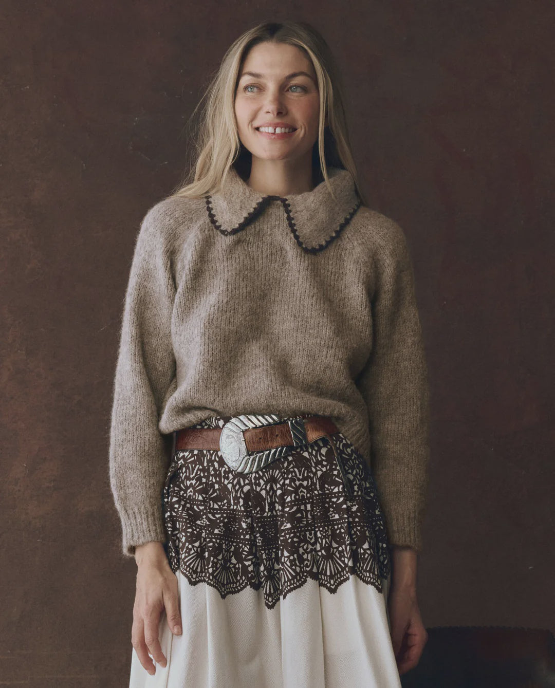 A woman with long blonde hair smiles while wearing The Great Inc.'s Crochet Collar Pullover—a brown knit sweater with a relaxed fit and scalloped crochet collar—paired with a wide belt and lace-patterned skirt, against a dark background.