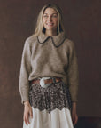 A woman with long blonde hair smiles while wearing The Great Inc.'s Crochet Collar Pullover—a brown knit sweater with a relaxed fit and scalloped crochet collar—paired with a wide belt and lace-patterned skirt, against a dark background.