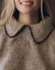 A woman with long blonde hair smiles, wearing The Great Inc.’s Crochet Collar Pullover—a relaxed fit beige sweater with a large rounded crochet collar and dark brown trim. Only her lower face and upper torso are shown.