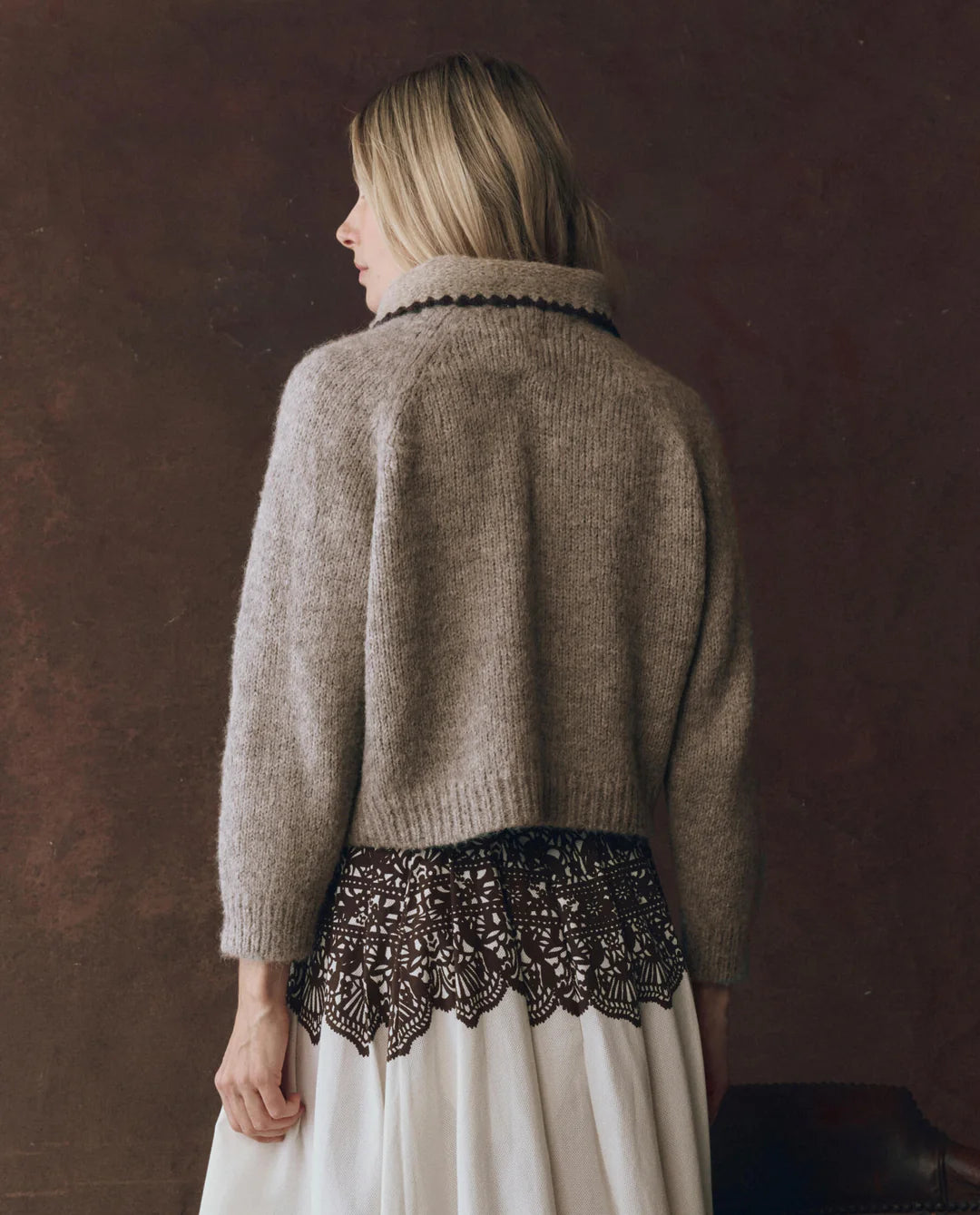 A woman with blonde hair is seen from behind, wearing The Crochet Collar Pullover by The Great Inc.—a cozy, relaxed fit light brown sweater—over a white dress with dark brown lace, set against a plain dark brown wall.