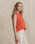A woman with wavy blonde hair models The Cashmere Pullover Vest by The Great Inc. in bright orange over a patterned collared shirt and white jeans, standing sideways against a textured neutral fabric background.