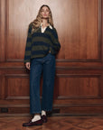 Academy Polo Sweater by The Great Inc. styled with wide-leg jeans and loafers beside wood-paneled wall.