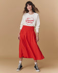 A woman poses against a beige background wearing The Great Inc. Greatest Mom Sweatshirt—white, 100% cotton with a red heart and "The Greatest mom"—paired with a red midi skirt, black high-top sneakers, and white socks.