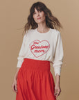A woman with wavy brown hair models The Great Inc.'s Greatest Mom Sweatshirt, featuring a red heart and "The Greatest Mom," paired with a high-waisted red skirt, standing against a plain beige background.
