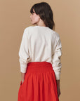 A person with long brown hair, seen from behind against a beige background, wears The Great Inc.'s cream-colored "Greatest Mom Sweatshirt" paired with a high-waisted bright red skirt.