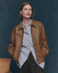 A woman with straight brown hair wears The Great Inc.’s vintage-inspired Flight Jacket over a striped button-up shirt and black pants, standing with her hands in her jacket pockets against a dark blue background.