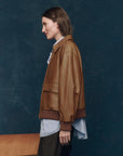 A woman with straight brown hair stands in profile against a dark blue background, wearing The Great Inc.'s The Flight Jacket—a vintage-inspired lamb leather piece—over a striped shirt and dark pants. A wooden surface is visible at the lower left.