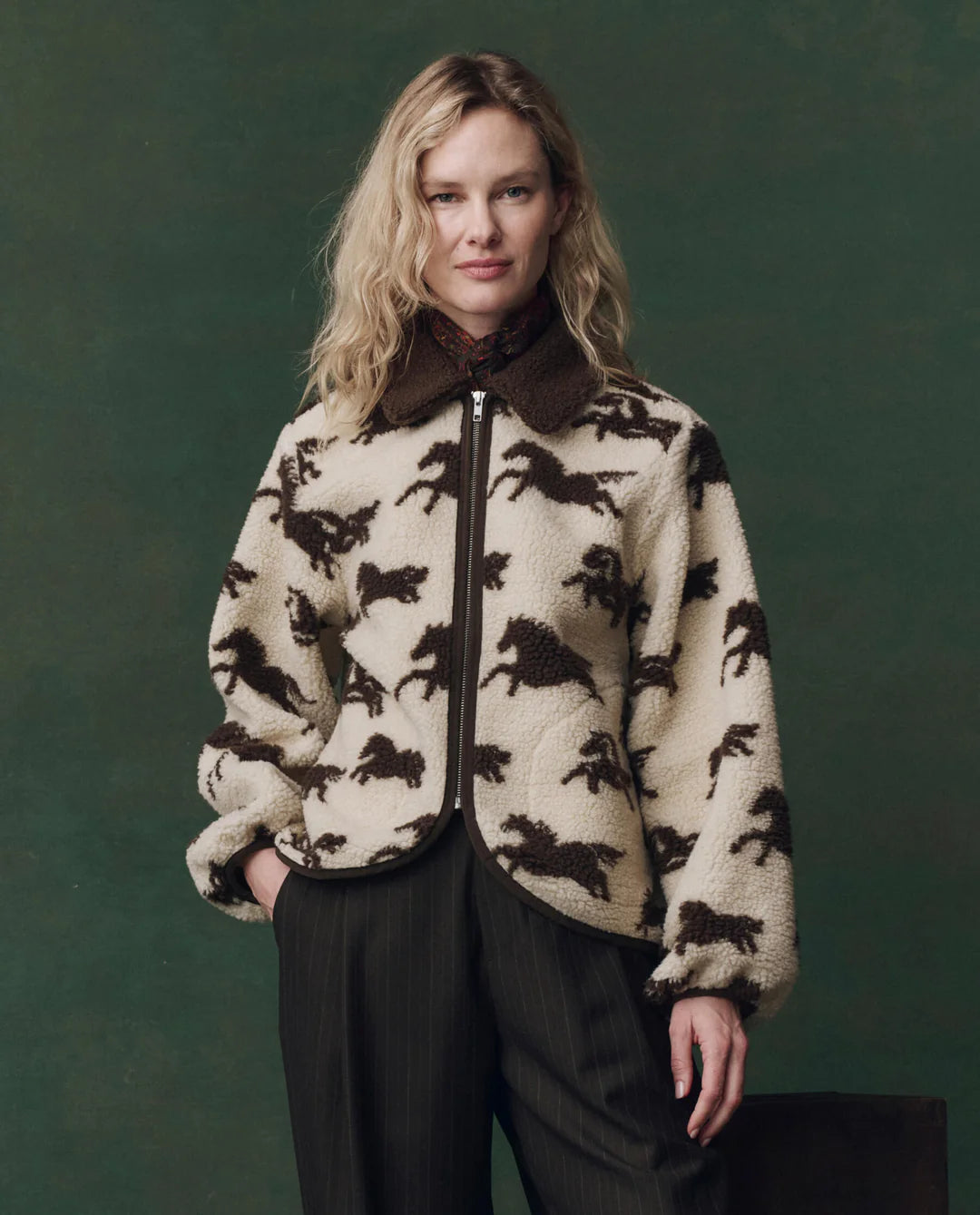 A woman with wavy blonde hair wears The Pasture Jacket by The Great Inc.—a cream teddy jacket with a horse motif, relaxed fit, dark collar, and wide sleeves—standing against a dark green background with a slight smile at the camera.