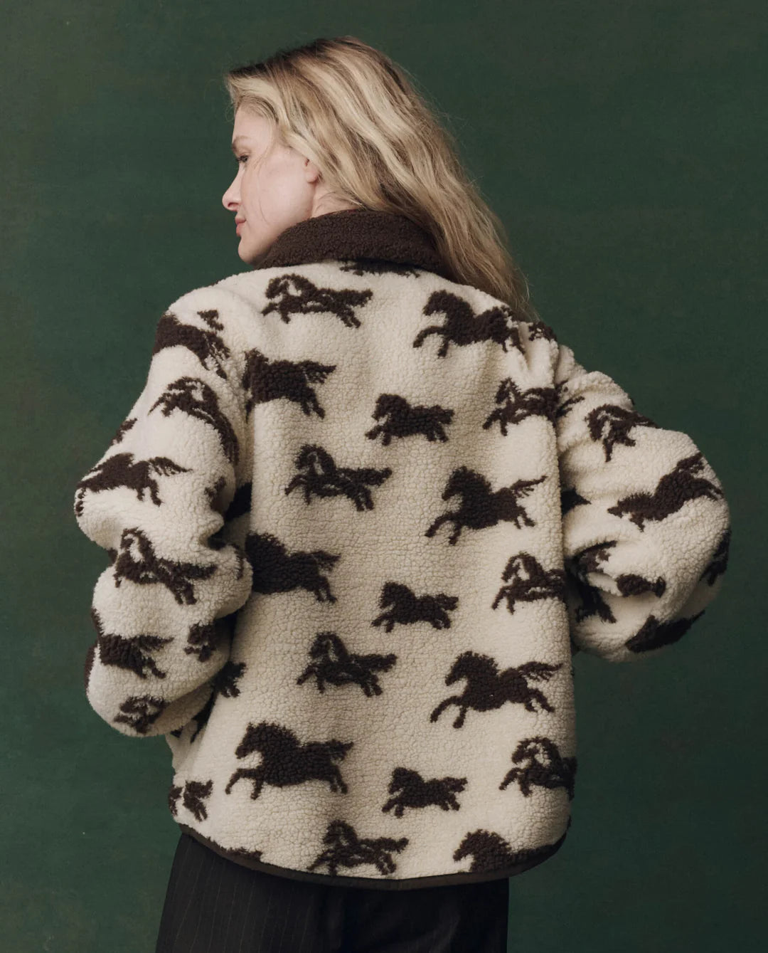 A person with long blonde hair stands sideways, wearing The Pasture Jacket by The Great Inc.—a relaxed fit teddy jacket featuring a brown horse motif—against a dark green background.