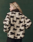 A person with long blonde hair stands sideways, wearing The Pasture Jacket by The Great Inc.—a relaxed fit teddy jacket featuring a brown horse motif—against a dark green background.