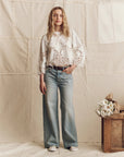 A woman stands before a neutral fabric backdrop, wearing The Picnic Top by The Great Inc., light-wash wide-leg jeans, and white sneakers. A wooden box with white flowers is positioned to her right.