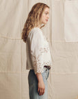 A woman with wavy blonde hair wears The Picnic Top by The Great Inc., featuring Battenberg lace sleeve details, paired with light blue jeans, standing in profile against a neutral quilted fabric background.