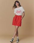 A woman stands against a beige background in The Great Inc.’s Greatest Mom Tee—featuring a red heart graphic—paired with red high-waisted shorts, white socks, and black sneakers. A perfect vintage-inspired look for fans of retro tees. Hands rest casually in her pockets.