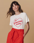 A woman with wavy brown hair wears The Great Inc. Greatest Mom Tee featuring "The Greatest mom" in a red heart, paired with high-waisted red pants, standing against a beige background.
