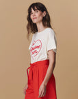 A woman with wavy brown hair wears The Great Inc. Greatest Mom Tee, a white jersey shirt with a red heart and "The Greatest Mom" text, paired with high-waisted bright red pants against a beige background.