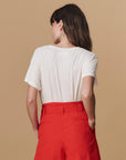 A woman with long brown hair faces away from the camera, wearing The Great Inc.'s Greatest Mom Tee, a white washed jersey t-shirt, paired with high-waisted bright red pants against a plain beige background.